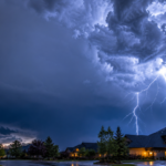 Photorealistic thunderstorm approaching a suburban neighborhood at dusk, dramatic lightning illuminating dark storm clouds, faint blue electrical glow in the air, cinematic atmosphere, highly detailed, realistic weather photography, mysterious mood. Large empty space in the sky for website title text. Colors: deep blues, grays, and white lightning.